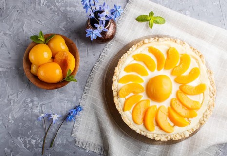 Round peach cheesecake and ceramic vase with blue flowers on a linen napkin on a gray concrete