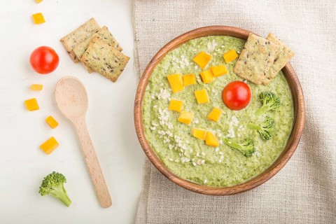 Green broccoli cream soup with crackers and cheese in wooden bowl on a white wooden background and