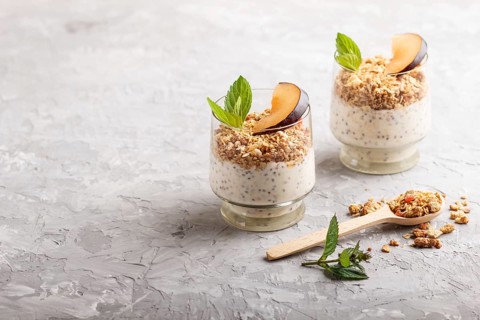 Yoghurt with plum, chia seeds and granola in a glass and wooden spoon on gray concrete background.