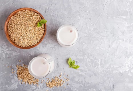 Organic non dairy oat milk in glass and wooden plate with oat seeds on a gray concrete background.
