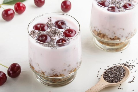Yoghurt with cherries, chia seeds and granola in glass with wooden spoon on white wooden background