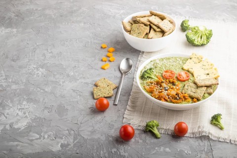 Green broccoli cream soup with crackers and cheese in white bowl on a gray concrete background and