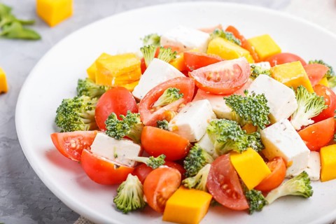 Vegetarian salad with broccoli, tomatoes, feta cheese, and pumpkin on white ceramic plate on a gray