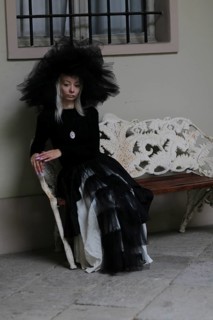 Gothically styled white thin blonde woman sitting on a lace-covered bench in a vintage indoor