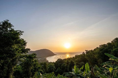 View from a mountain onto a secluded bay with a sandy beach and mangrove forest. The sun sets over