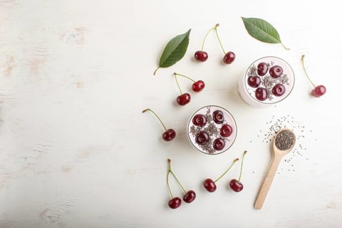Yoghurt with cherries, chia seeds and granola in glass with wooden spoon on white wooden background