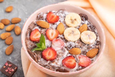 Chocolate cornflakes with milk, strawberry and almonds in ceramic bowl on gray concrete background