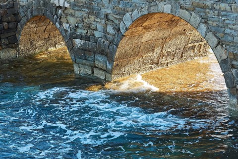 Sunlight shines through stone arches of an old bridge with foaming sea waves below, sea fortress