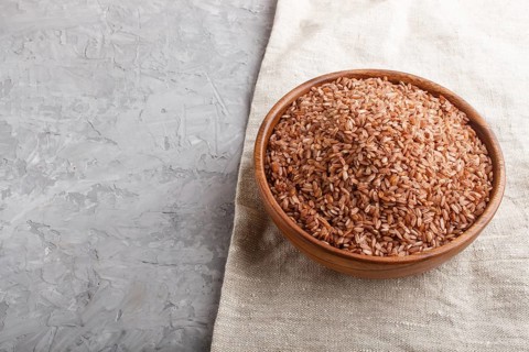 Wooden bowl with unpolished brown rice on a gray concrete background and linen textile. Side view,