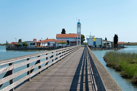 A wooden footbridge leads over calm waters to a monastery under a clear blue sky, Monastery of St