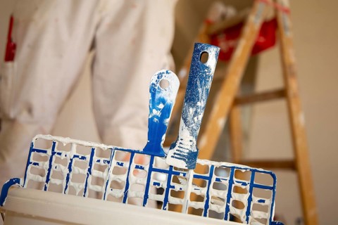 Symbolic image of painting work: Close-up of a paint bucket with scraper grid, in the background a