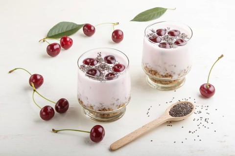 Yoghurt with cherries, chia seeds and granola in glass with wooden spoon on white wooden background