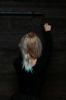Back view of a blonde thin white woman dressed in black reaching up to a rusty antique metal lock