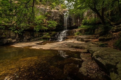 A secluded waterfall spills over ancient rocks into a still pool, shaded by a wooded canopy, in