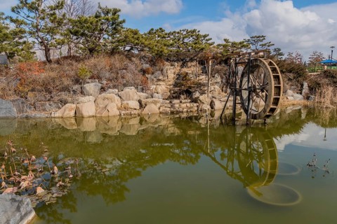 A water wheel beside a peaceful pond with rock formations and clear reflections, in South Korea