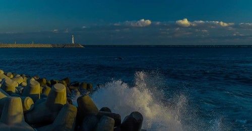 Evening view of a calm seashore with spray over tetrapods, lighthouse in the background, in South