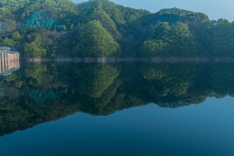Calm reservoir with forested hills and morning mist, creating a mirror-like reflection, in South