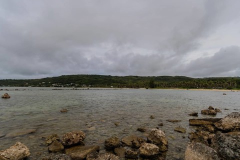 Seascape of rocky shoreline on a cloudy day with buildings on tree lined shore in the distance in