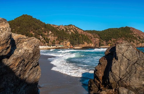 The ocean's waves crash against a cliffside with forested hills under the clear sky, in South Korea