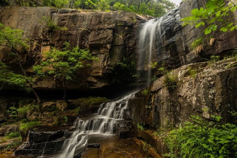 Sunlight filters through the trees onto a cliff from which a waterfall descends into a rocky basin,