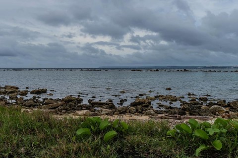 Seascape of rocky shoreline on a cloudy day with buildings on tree lined shore in the distance in