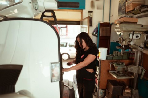 Mechanic standing by a classic scooter in a sunlit workshop full of tools, latino female in