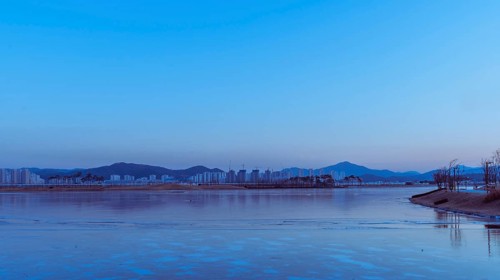 Twilight cityscape with a calm river partially covered in ice, reflecting blue hues, in South Korea