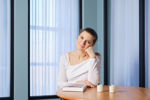 Dreamy woman sits at a wooden table with a book, props her head up with her fist and looks with a