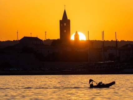 Motorboat in the golden evening light at sunset, silhouette of the church towers of Rab, town of
