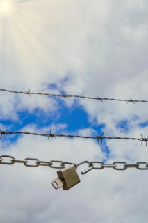 Chain with padlock open with key next to a barbed wire with a blue cloudy sky with sun rays,
