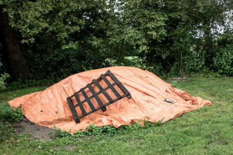 An orange tarpaulin covers something on the ground next to a wooden pallet and bushes