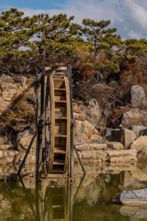 Close-up of a water wheel with its reflection in a pond, set against a rocky backdrop, in South