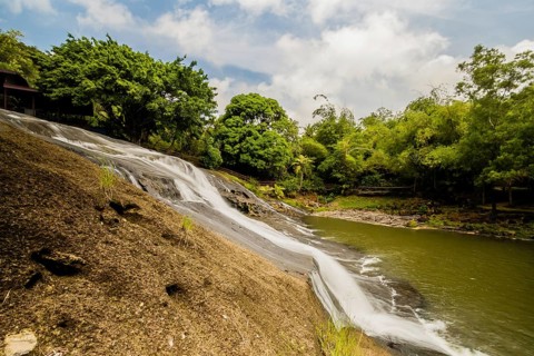 Side view of a waterfall cascading down a cliff into the basin below with green trees and blue,