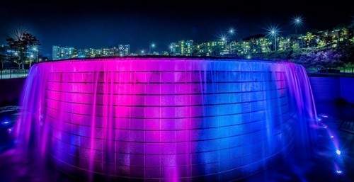 Night scene of waterfall lit up with blue and red lights with the lights of buildings of a town in
