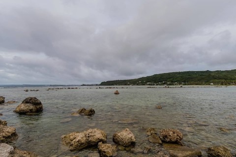 Seascape of rocky shoreline on a cloudy day with buildings on tree lined shore in the distance in