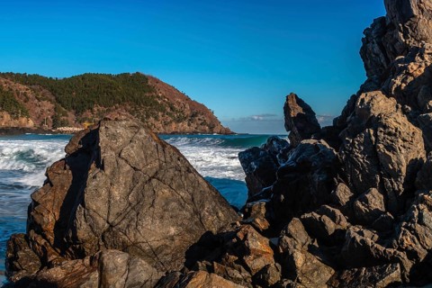 Frothy ocean waves kiss the rocky landscape under the expansive blue skies, in South Korea