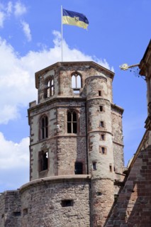 The top of a historic tower with flag and battlements (Heidelberg Castle), under a blue sky,