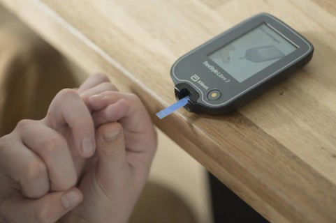 Blood glucose meter with test strips lying on a wooden table, child's hand holding a finger and