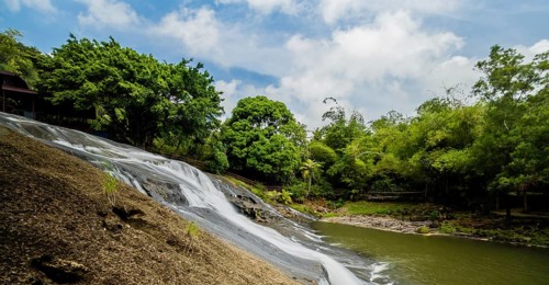 Side view of a waterfall cascading down a cliff into the basin below with green trees and blue,