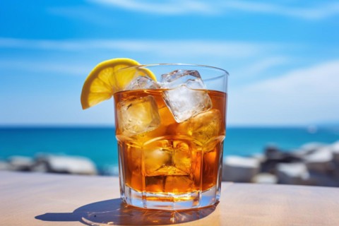Drinking glass with cocktail, lemon slice and ice cubes with blurry ocean in background. KI