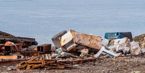 Old rusted metal refrigerator along with other trash and debris laying on the ground with the ocean