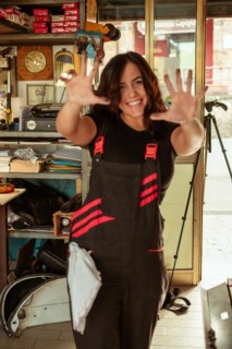 Cheerful hispanic young female mechanic wearing black overalls waving her hands in a workshop,