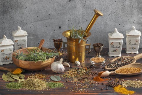 Antique spice containers and a mortar surrounded by fresh herbs and spices on a wooden surface