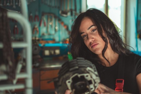Woman mechanic holding a motorcycle engine part in a workshop, appearing thoughtful, a complete