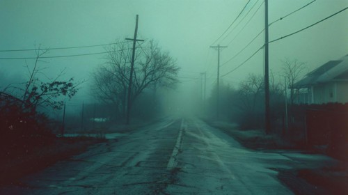 A desolate road stretches into fog, lined by power lines and silhouettes of houses, AI generated
