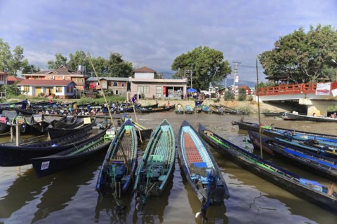 View of calm waterway with anchored boats, a bridge in the background and surrounding houses, Inle