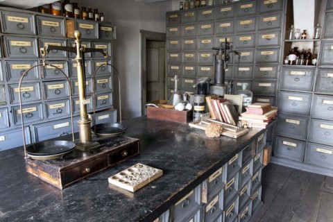 A pharmacist's scales stand on a worktable in front of shelves with drawers containing ingredients
