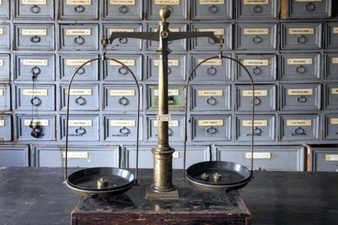 A pharmacist's scales stand on a worktable in front of shelves with drawers containing ingredients