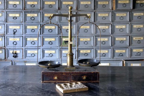 A pharmacist's scales stand on a worktable in front of shelves with drawers containing ingredients