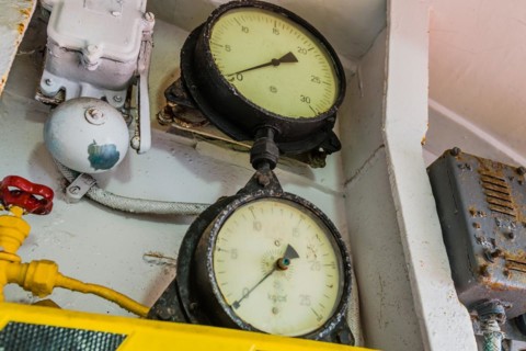 Old pressure gauges mounted above yellow and black striped ledge with valves and bell in background